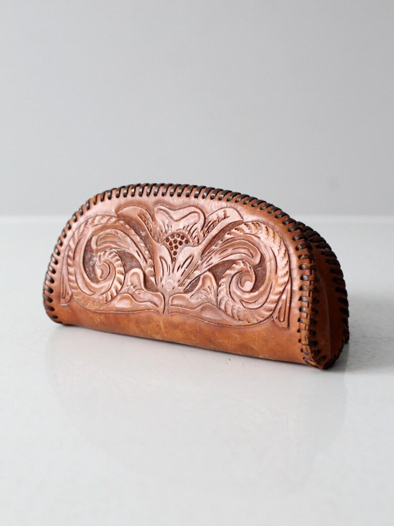 May include: A brown leather clutch purse with a floral design embossed into the leather. The purse has a curved shape and a stitched edge.