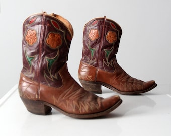 May include: A pair of brown leather cowboy boots with floral embroidery in shades of orange, green, and purple. The boots have pointed toes and a slight heel.