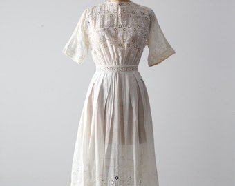 May include: A vintage white dress with short sleeves and a high neckline. The dress features intricate eyelet embroidery on the bodice, sleeves, and skirt. The waist is cinched with a delicate band, and the skirt falls to a mid-calf length.