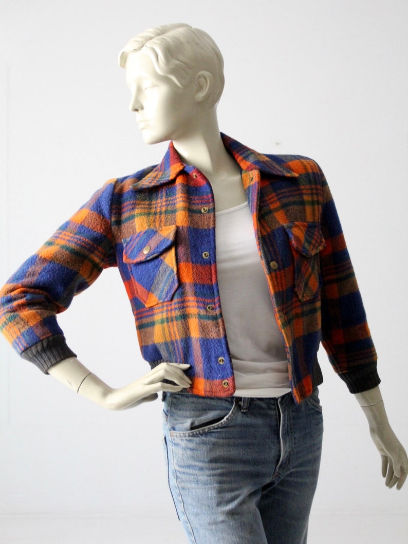 May include: A blue, orange, and brown plaid flannel jacket with a button-up front and two flap pockets. The jacket has a cropped length and a ribbed knit trim at the cuffs and hem.