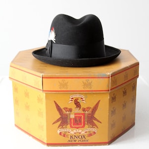 Op de afbeelding: Een zwarte fedora hoed met een zwart lint en een decoratieve veer staat op een vintage gele hoedendoos. De hoedendoos heeft een wapen met de woorden "Knox New York".