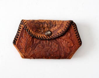 May include: A small, brown leather coin purse with a floral embossed design. The purse has a snap closure and a decorative stitch around the edge.