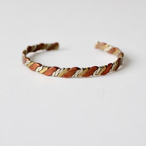Op de afbeelding: Een gevlochten koperen, zilveren en gouden armband. De armband is gemaakt van drie verschillende metalen die samen zijn geweven om een uniek en stijlvol ontwerp te creëren.