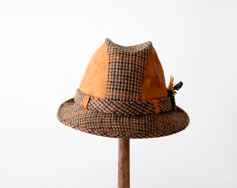 May include: A brown and tan tweed fedora hat with a suede crown and a small feather accent. The hat is displayed on a wooden stand.