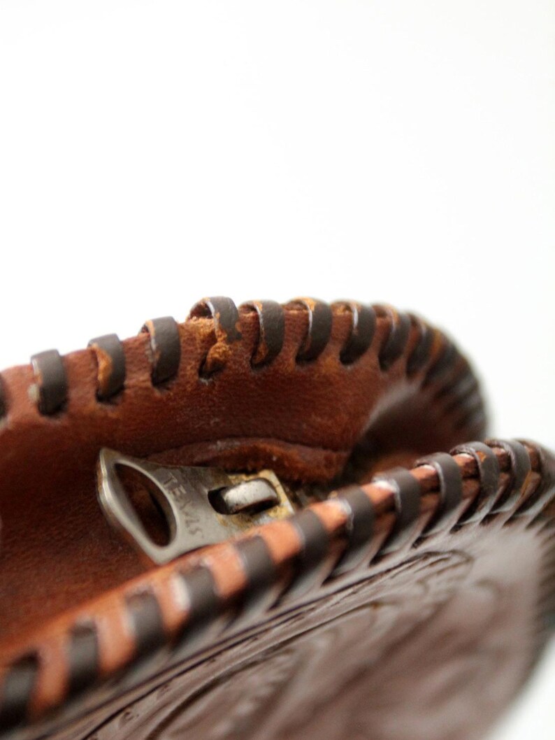 May include: Close-up of a brown leather coin purse with a zipper closure. The purse has a decorative stitched design around the edge.
