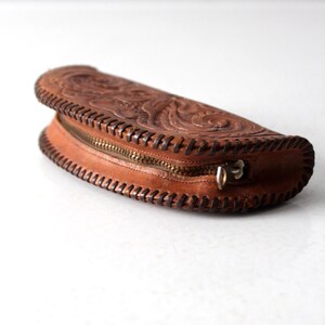 May include: A brown leather clutch with intricate floral tooling and a zipper closure. The clutch is shaped like a half-moon and has a stitched edge.