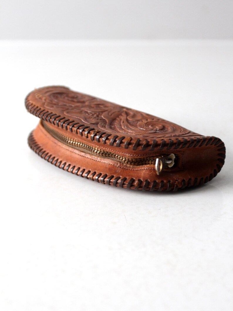 May include: A brown leather clutch with intricate floral tooling and a zipper closure. The clutch is shaped like a half-moon and has a stitched edge.