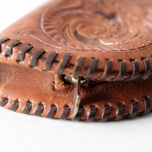 May include: Close-up of a brown leather purse with intricate embossed floral designs. The purse has a zipper closure and a dark brown stitched edge.