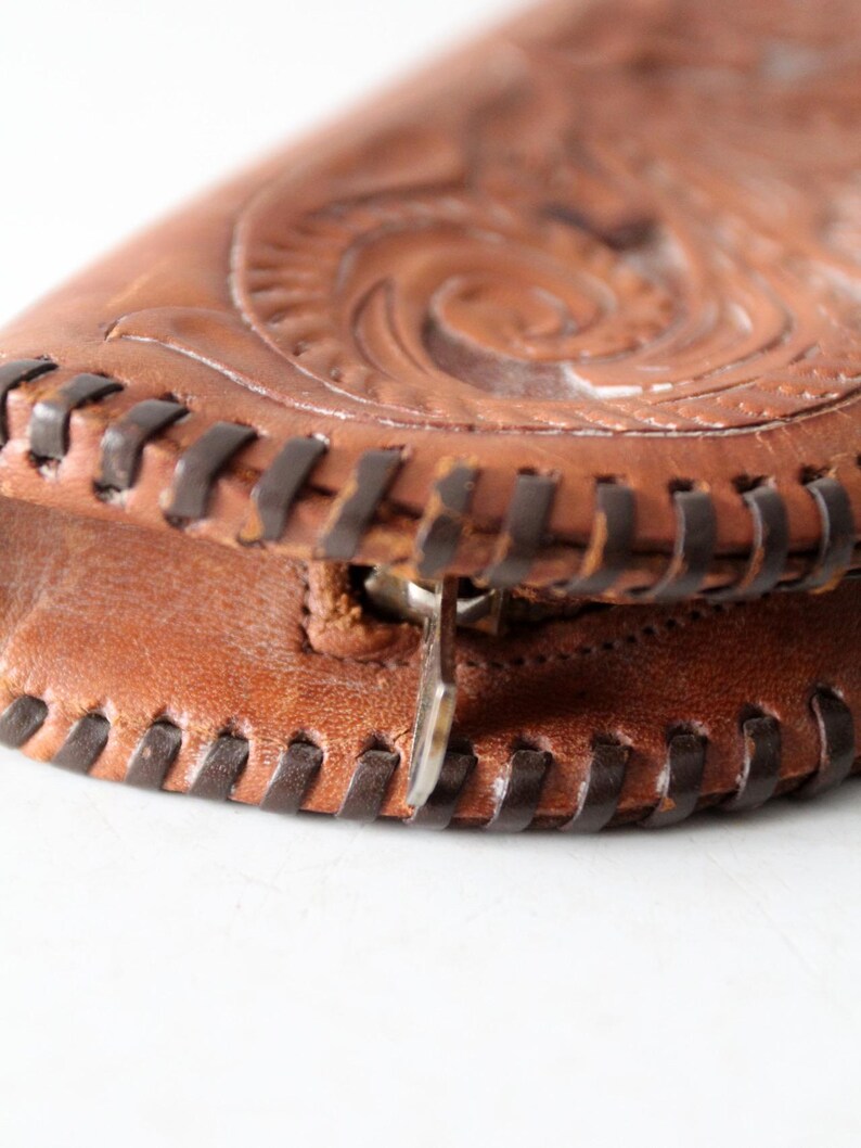 May include: Close-up of a brown leather purse with intricate embossed floral designs. The purse has a zipper closure and a dark brown stitched edge.
