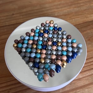 May include: A square-shaped grid of small, spherical beads in various colours including metallic gold, silver, shades of blue, brown, and beige. The beads are strung together in a dense, interconnected pattern, creating a textured, multi-coloured surface.
