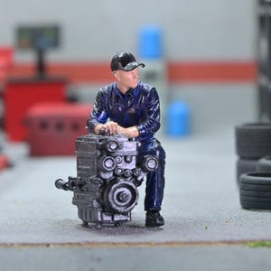 May include: A miniature figurine of a mechanic in a blue uniform and black cap, leaning against a detailed engine. The scene is set in a garage with tires stacked in the background. The engine is a dark gray color.