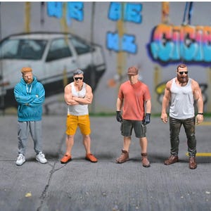 May include: Four miniature male figurines in casual clothes stand on a grey surface in front of a graffiti-covered wall. The figures wear hoodies, vests, shorts, and sunglasses. The background includes a car and colourful graffiti.