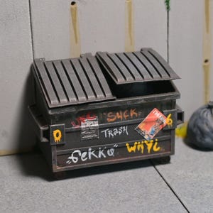May include: A miniature, weathered dumpster with graffiti, including the words "Suck Trash" and "Sekko Whyi." A crumpled gray trash bag sits nearby. The dumpster's lid is partially open.