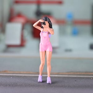 May include: Miniature figure of a person in a pink jumpsuit, holding a camera. The figure is wearing purple sneakers and a light purple top. The background is blurred, suggesting an outdoor or industrial setting. The figure is posed as if taking a photo.