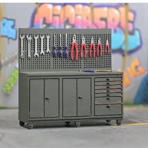 May include: A miniature gray workshop with a pegboard back, featuring various tools like wrenches, screwdrivers, and pliers. The workshop has cabinets, drawers, and is set on a gray surface. The background includes graffiti art.