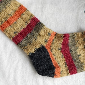 May include: Hand-knitted sock in progress, featuring a color-blocked design. The sock showcases stripes of orange, red, and tan, with a dark gray heel and toe. Knitting needles and yarn are visible, indicating a craft project.