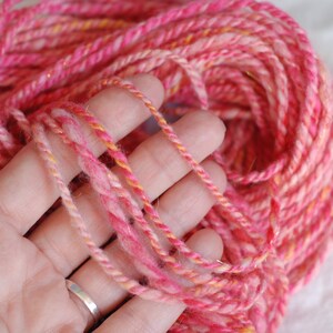 May include: Pink and orange yarn with a subtle shimmer, held in a hand. The yarn is soft and fluffy, perfect for knitting or crocheting.