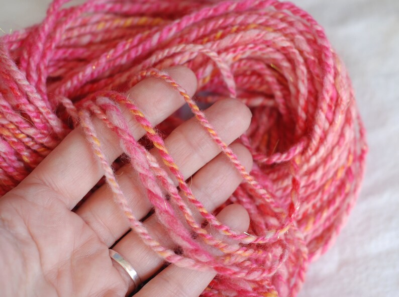 May include: Pink and orange yarn with a subtle shimmer, held in a hand. The yarn is soft and fluffy, perfect for knitting or crocheting.