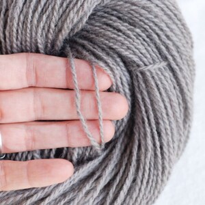 Puede incluir: Un ovillo de lana gris, sostenido en una mano, que muestra la textura y el color de la lana.