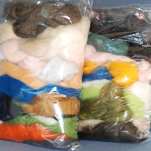 May include: Two clear plastic bags filled with various colours of wool roving. The bags are laid out on a blue fabric surface. The wool roving is arranged in layers of different colours, including brown, white, pink, blue, green, yellow, and orange.