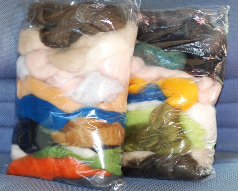 May include: Two clear plastic bags filled with various colours of wool roving. The bags are laid out on a blue fabric surface. The wool roving is arranged in layers of different colours, including brown, white, pink, blue, green, yellow, and orange.