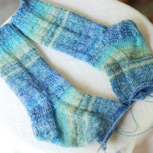 May include: A pair of hand-knitted socks in shades of blue, turquoise, and pale yellow. The socks are made of soft yarn and are partially finished, with needles and yarn visible. The color gradient creates a watercolor effect.