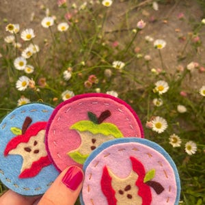 May include: Three felt apple-shaped patches in blue, pink, and light pink, each with a different apple design. The patches are hand-stitched with contrasting thread and feature red, green, and tan apple details. The background is a field of daisies.