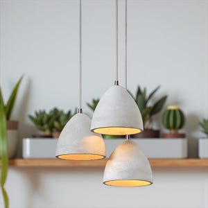 Puede incluir: Tres lámparas colgantes con un acabado texturizado blanco roto cuelgan del techo. Las lámparas tienen un diseño en forma de campana con un brillo cálido. El fondo presenta una estantería de madera con plantas en macetas.