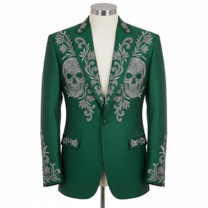 May include: A green blazer with silver skull and floral embroidery. The jacket has detailed embroidery on the lapels, shoulders, and sleeves. The blazer is displayed on a mannequin, showcasing its tailored fit and design.