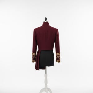 K&ouml;nnte beinhalten: Burgunderfarbene Jacke mit goldfarbenen Stickereien an den &Auml;rmeln. Die Jacke wird auf einer schwarzen Schneiderpuppe mit wei&szlig;em St&auml;nder pr&auml;sentiert. Die Jacke hat einen hohen Kragen und einen drapierten Einsatz auf der linken Seite.