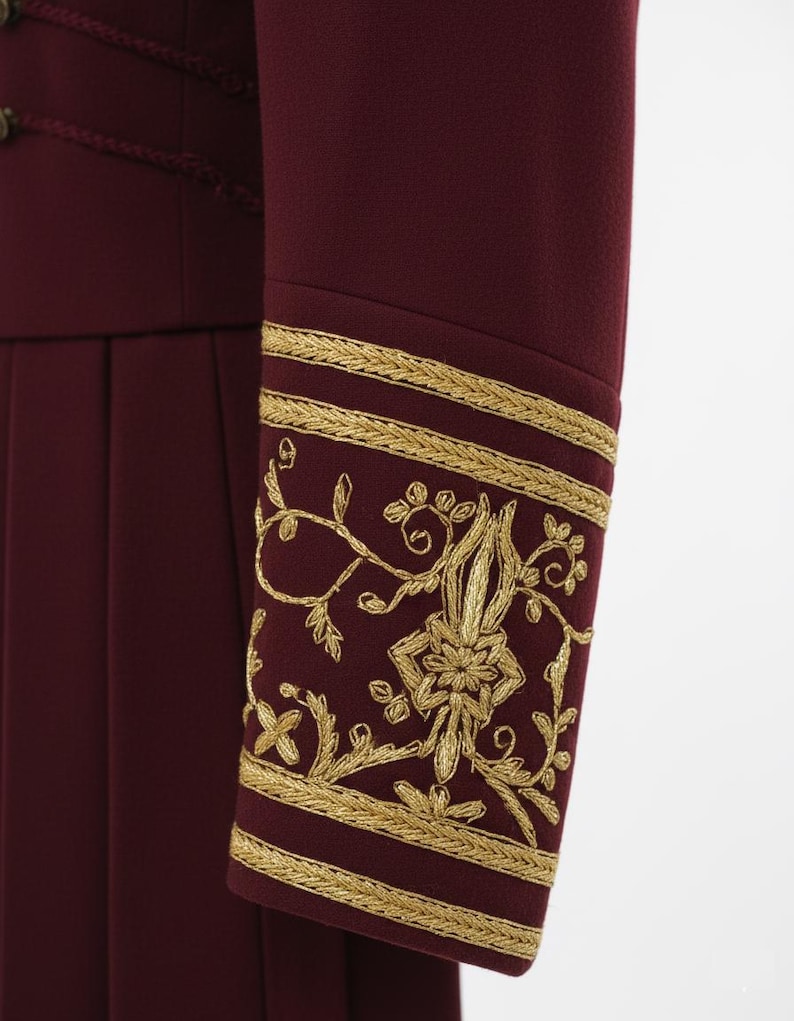 K&ouml;nnte beinhalten: Nahaufnahme eines bordeauxfarbenen &Auml;rmels mit goldener Stickerei. Der &Auml;rmel zeigt detaillierte goldene Blumen- und Blattmuster mit goldenen Zierleisten. Das Kleidungsst&uuml;ck hat ein plissiertes Design.