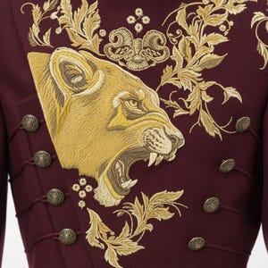 K&ouml;nnte beinhalten: Ein burgunderfarbener Mantel mit aufwendiger goldener Stickerei, die einen br&uuml;llenden L&ouml;wenkopf und florale Muster zeigt. Der Mantel hat dekorative Kn&ouml;pfe und einen hohen Kragen und zeigt ein k&ouml;nigliches und detailliertes Design.