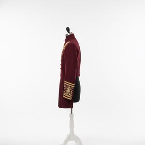 K&ouml;nnte beinhalten: Burgunderfarbene Jacke mit goldener Stickerei auf Schultern und Manschetten, pr&auml;sentiert auf einer wei&szlig;en Schaufensterpuppe. Die Jacke hat einen hohen Kragen und goldene Verzierungen an den &Auml;rmeln. Die Schaufensterpuppe steht auf einem wei&szlig;en St&auml;nder.