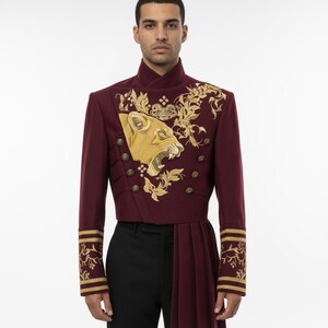 K&ouml;nnte beinhalten: Burgunderfarbene Jacke im Milit&auml;rstil mit goldener Stickerei eines L&ouml;wenkopfes und floralen Designs. Die Jacke hat goldene Kn&ouml;pfe, einen hohen Kragen und plissierte Stoffdetails. Die &Auml;rmel haben goldene Verzierungen. Die Jacke wird mit einer schwarzen Hose getragen.
