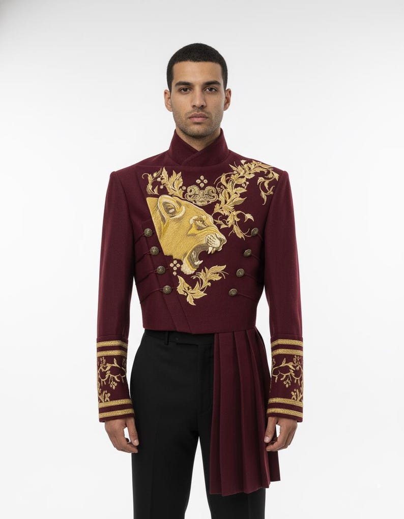K&ouml;nnte beinhalten: Burgunderfarbene Jacke im Milit&auml;rstil mit goldener Stickerei eines L&ouml;wenkopfes und floralen Designs. Die Jacke hat goldene Kn&ouml;pfe, einen hohen Kragen und plissierte Stoffdetails. Die &Auml;rmel haben goldene Verzierungen. Die Jacke wird mit einer schwarzen Hose getragen.