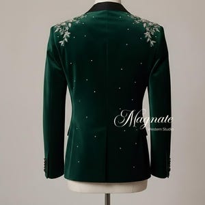 Puede incluir: Una blazer de terciopelo verde oscuro con solapa negra y bordado floral plateado en los hombros. La blazer presenta detalles de pedrer&iacute;a dispersos y la palabra "Magnate" en escritura cursiva.