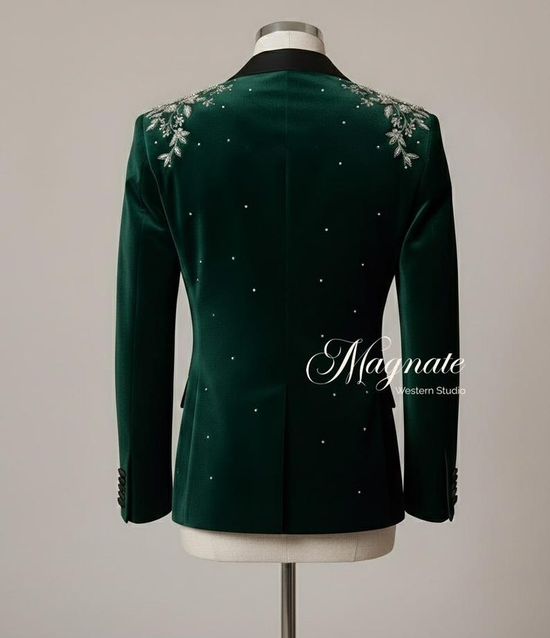 Puede incluir: Una blazer de terciopelo verde oscuro con solapa negra y bordado floral plateado en los hombros. La blazer presenta detalles de pedrer&iacute;a dispersos y la palabra "Magnate" en escritura cursiva.