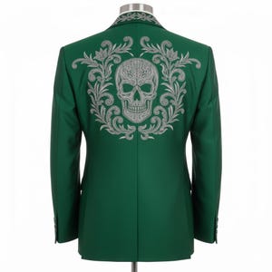 May include: A dark green blazer featuring a silver embroidered skull and floral design on the back. The blazer is tailored and displayed on a mannequin. The design is intricate and detailed, with a classic fit.