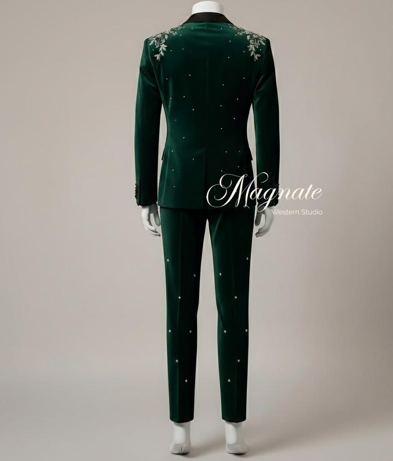 Puede incluir: Un traje de terciopelo verde oscuro con solapa negra, adornado con bordados florales plateados en los hombros y pedrer&iacute;a dispersa. El traje incluye una chaqueta y un pantal&oacute;n a juego. El texto "Magnate Western Studio" es visible.