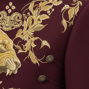 K&ouml;nnte beinhalten: Burgunderfarbene Jacke mit aufwendiger goldener Stickerei eines L&ouml;wenkopfes und floralen Mustern. Sie verf&uuml;gt &uuml;ber verzierte goldene Kn&ouml;pfe und Kordelverschl&uuml;sse. Das Design der Jacke ist detailliert und luxuri&ouml;s, geeignet f&uuml;r formelle Anl&auml;sse.