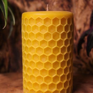 Puede incluir: Una vela cilíndrica de cera de abeja amarilla con un patrón de panal. La vela tiene una mecha y está colocada sobre un fondo de madera borroso. El diseño de la vela es un patrón repetitivo de celdas hexagonales.