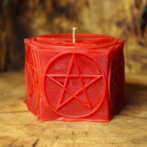 Può includere: Una candela rossa a forma di cubo con un disegno a pentagramma. La candela ha uno stoppino in cima ed è decorata con pentagrammi all'interno di cerchi su ogni lato. La candela è su una superficie di legno.
