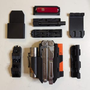 Pode incluir: Uma variedade de acessórios pretos para multi-ferramentas e uma multi-ferramenta Leatherman Wave. Os acessórios incluem uma faca suíça vermelha, uma lanterna preta e vários porta-bits. A Leatherman Wave é prateada e preta.