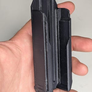 May include: A black, rectangular 3D-printed object with a unique design, held in a hand. The object has a series of vertical grooves and a distinct shape, possibly a magazine holder or similar accessory.