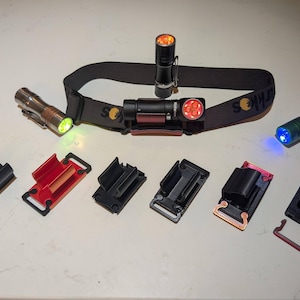 May include: A selection of flashlights and accessories. The image shows a black headlamp with a red light, several handheld flashlights, and various black and red mounting clips. The flashlights emit green, orange, and blue light.