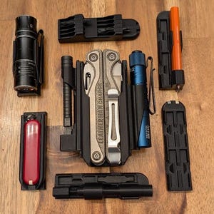 May include: A collection of black multi-tool holders and accessories, including a Leatherman multi-tool, flashlights, and a red knife. The items are arranged on a wooden surface.