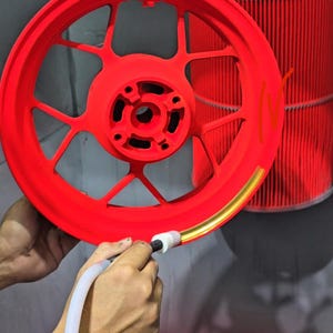 May include: A bright red motorcycle wheel rim with a multi-spoke design being painted with a gold metallic stripe along the outer edge using a handheld applicator tool. The wheel features a central hub with bolt holes and a matte finish.