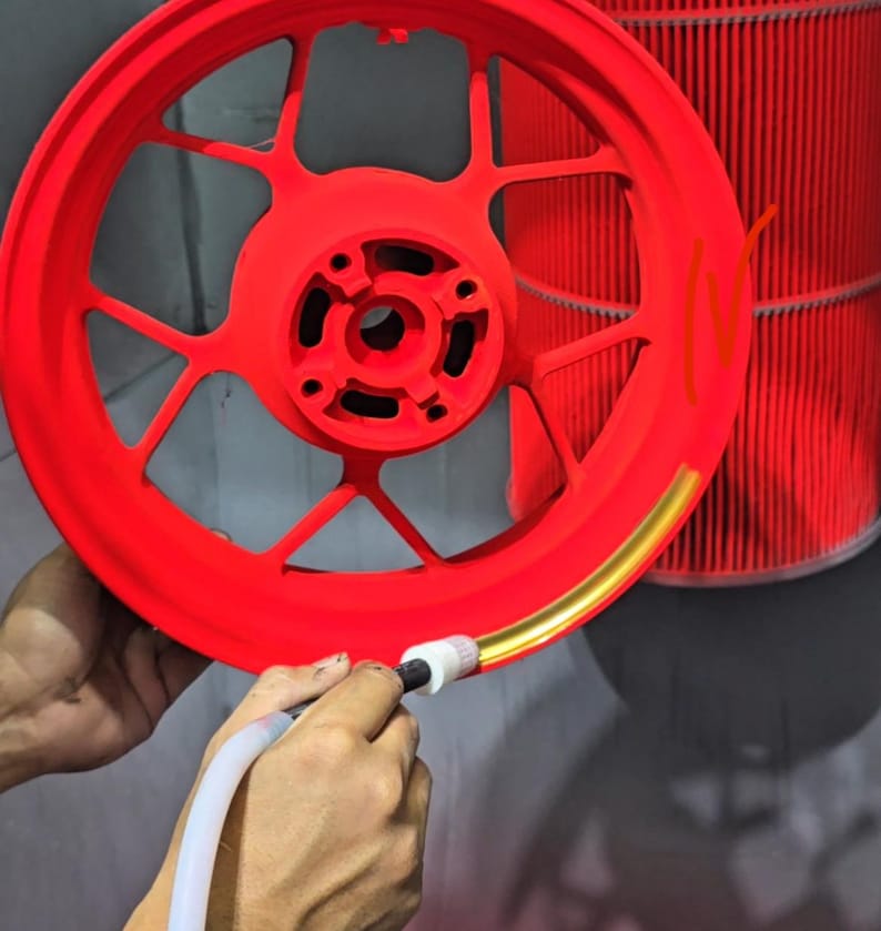 May include: A bright red motorcycle wheel rim with a multi-spoke design being painted with a gold metallic stripe along the outer edge using a handheld applicator tool. The wheel features a central hub with bolt holes and a matte finish.