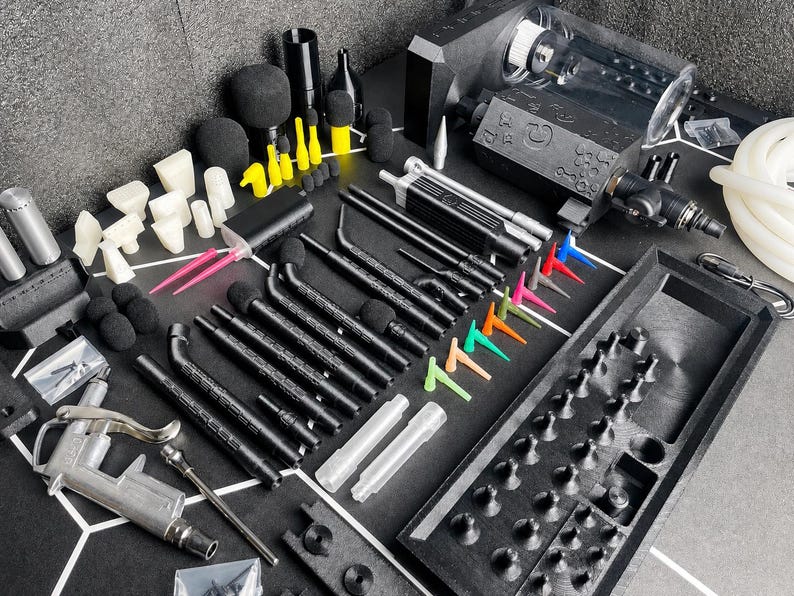 May include: A comprehensive set of black and silver automotive detailing tools including a pneumatic blow gun, various black extension wands with foam tips, colorful plastic nozzles, a black storage tray, and a white hose on a black surface.