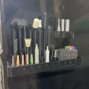 May include: A black plastic organizer shelf mounted on a dark surface, featuring multiple vertical slots holding various black and silver tools, small colorful nozzles, and a textured gray foam block for storing additional small components.